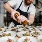 Brodaty kucharz doprawia pieprzem danie w ruchliwej kuchni restauracyjnej